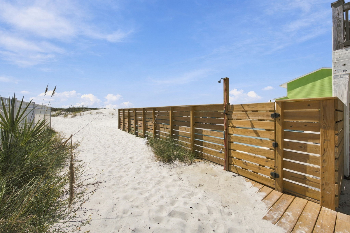 Sandy walkway beside the coastal vacation home leading toward the beach, surrounded by native beach vegetation and privacy fencing.