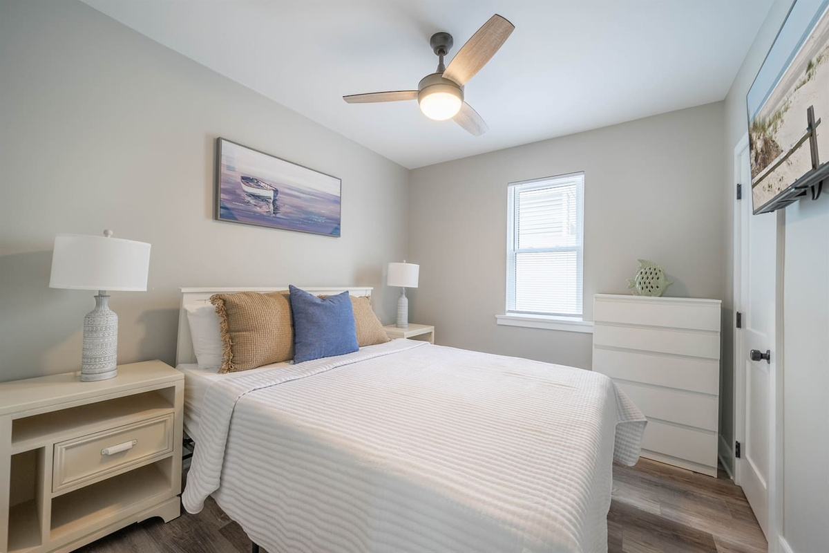 Bright bedroom with a comfortable bed, soft linens, and coastal-inspired decor. A ceiling fan, bedside lighting, and simple furnishings create a calm space for restful nights, with natural light adding to the relaxed atmosphere.