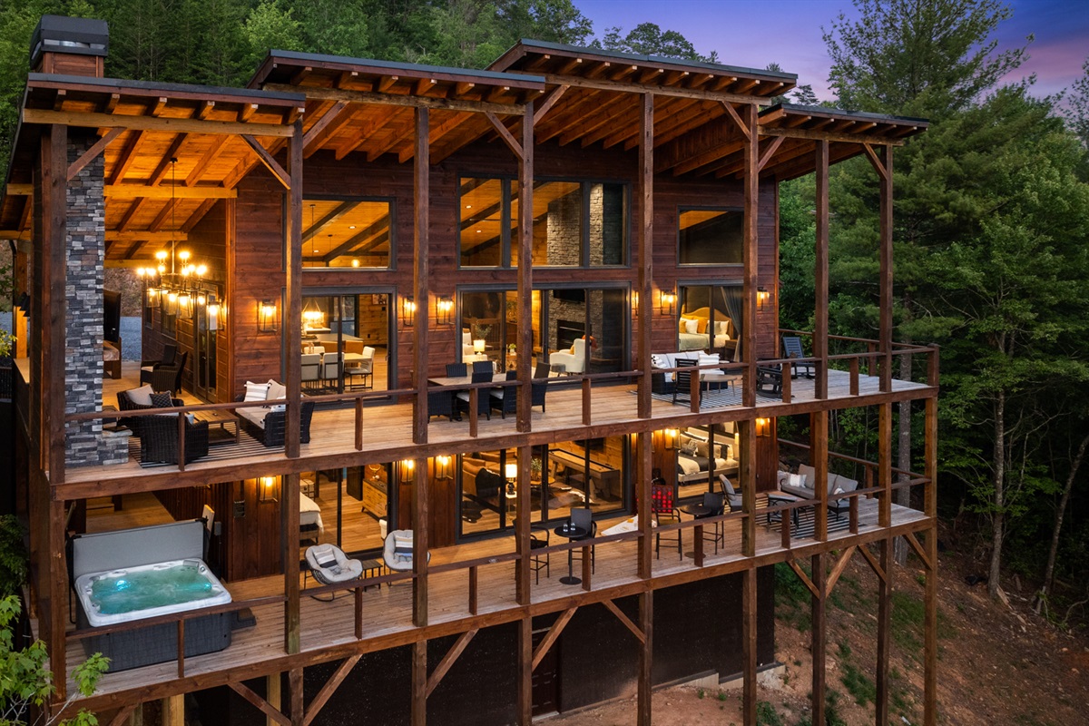 Expansive glass walls, warm ambient lighting, multiple decks with luxurious seating, a hot tub, and breathtaking forest views.