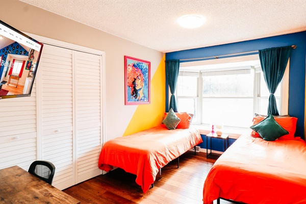 Twin Tranquility: Embrace Comfort in this Refreshing Bedroom with Twin Beds, Bathed in Vibrant Orange Hues and Complemented by Cool Bluish-Green Curtains. Enjoy the View and Entertainment with a Window and TV Screen 🛏️🪟📺