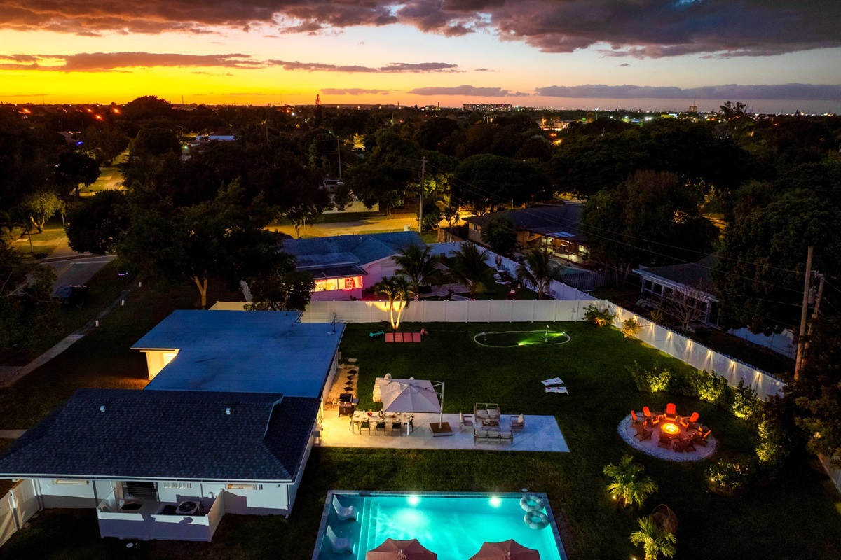 Private Palm Beach backyard at sunset with illuminated pool, fire pit seating, and landscaped grounds.