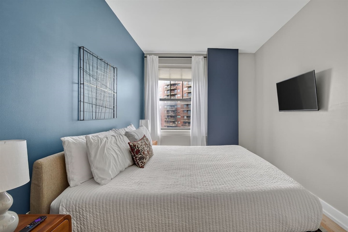 Relax in a serene bedroom featuring a plush king bed, crisp linens, smart TV, and bold accent wall. Wake up to natural light and city views—perfect for restful nights and refreshed mornings in downtown Des Moines.