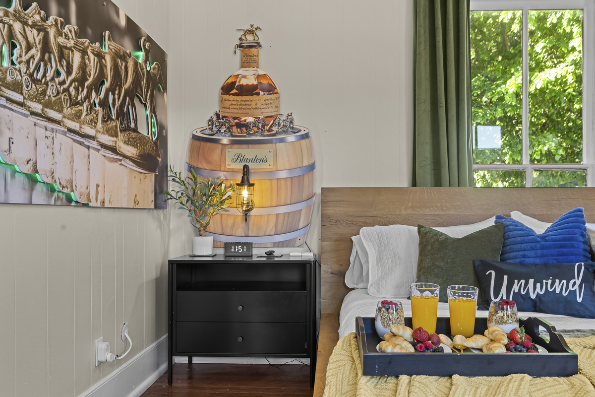 Green curtains, a Blanton’s barrel lamp, and bedside reading within reach make for a well-considered nightstand in a thoughtfully designed room.