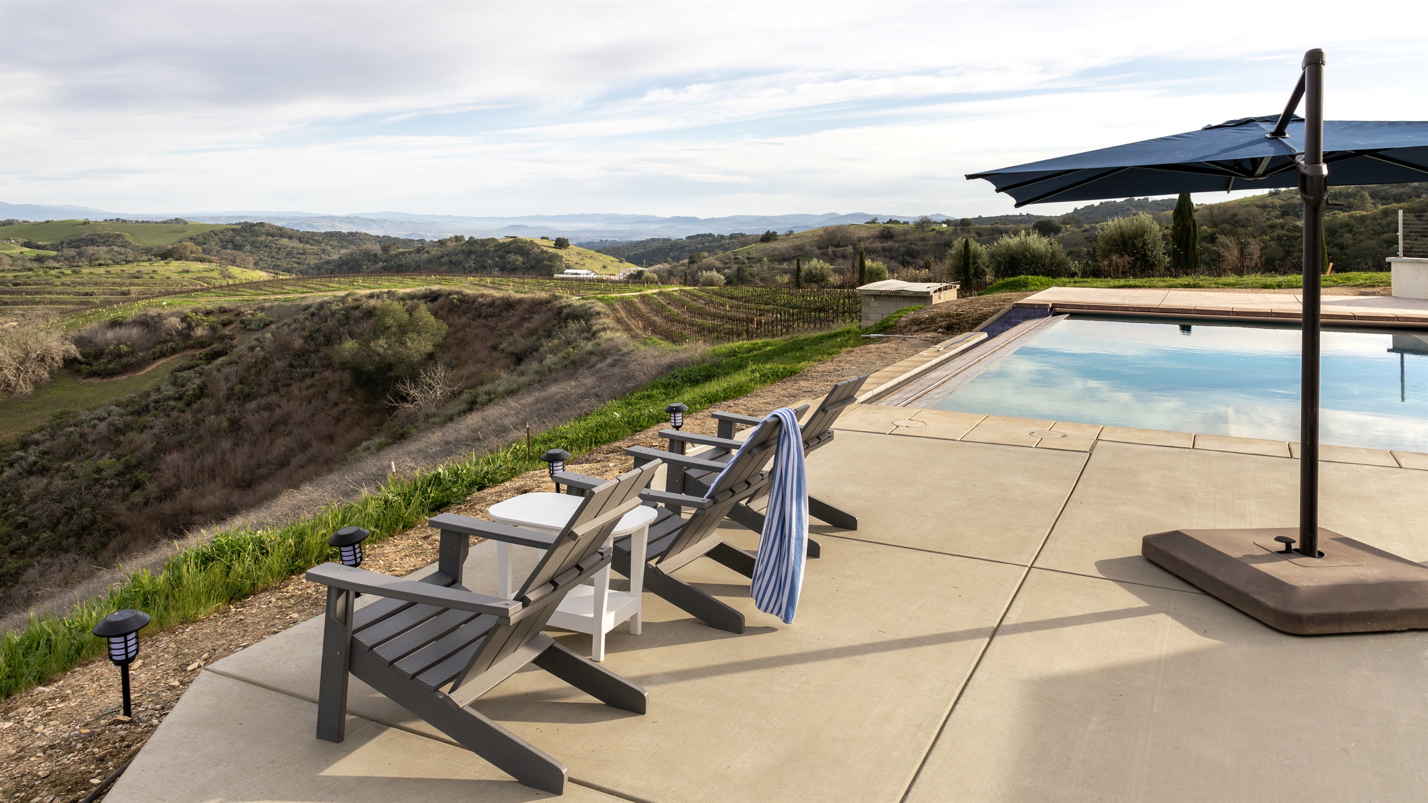 The full picture — cushioned loungers by the pool, the outdoor dining table beyond, Adirondack chairs at the infinity edge, and miles of wine country in every direction. Everything you need, nothing you don't.