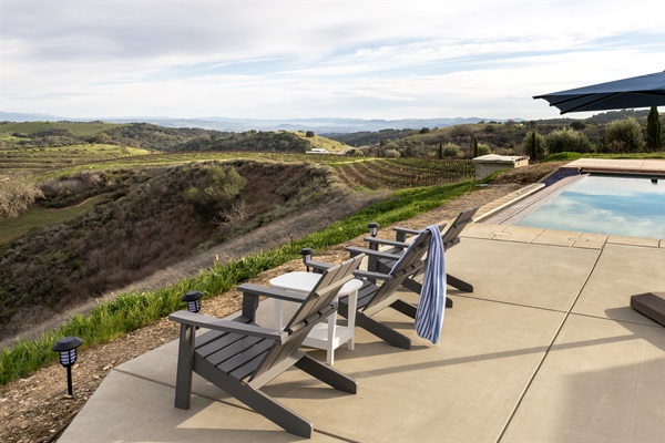The full picture — cushioned loungers by the pool, the outdoor dining table beyond, Adirondack chairs at the infinity edge, and miles of wine country in every direction. Everything you need, nothing you don't.
