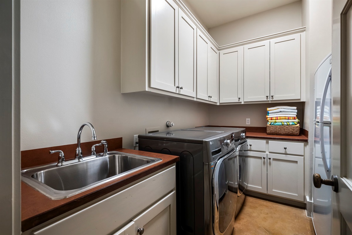 A well-equipped laundry room featuring a stainless steel utility sink, modern front-loading washer and dryer, white cabinetry for storage, and a countertop with stacked, colorful towels.
