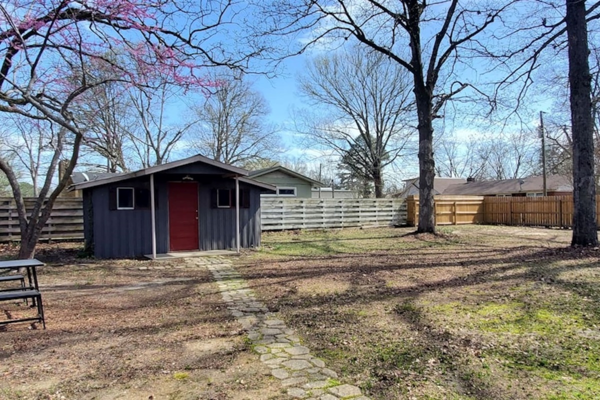 Spacious backyard with a cozy gray shed, red door, and stone path. Surrounded by trees and fencing—perfect for relaxing or entertaining.