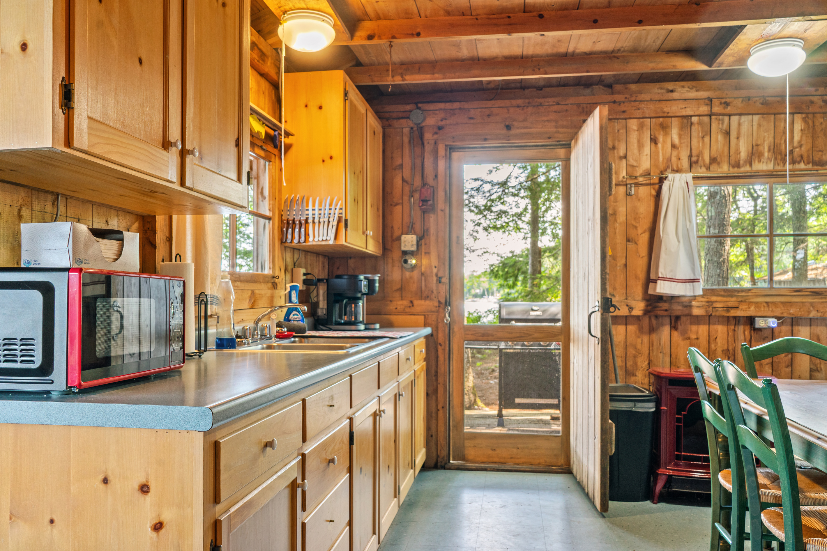 Kitchen counter workspace at Loonrise Point! Features microwave, drip coffee maker, knife block, and double sink. Door opens toward the outdoor grill with easy access to the red heater stove on Beech Hill Pond in Otis, ME.