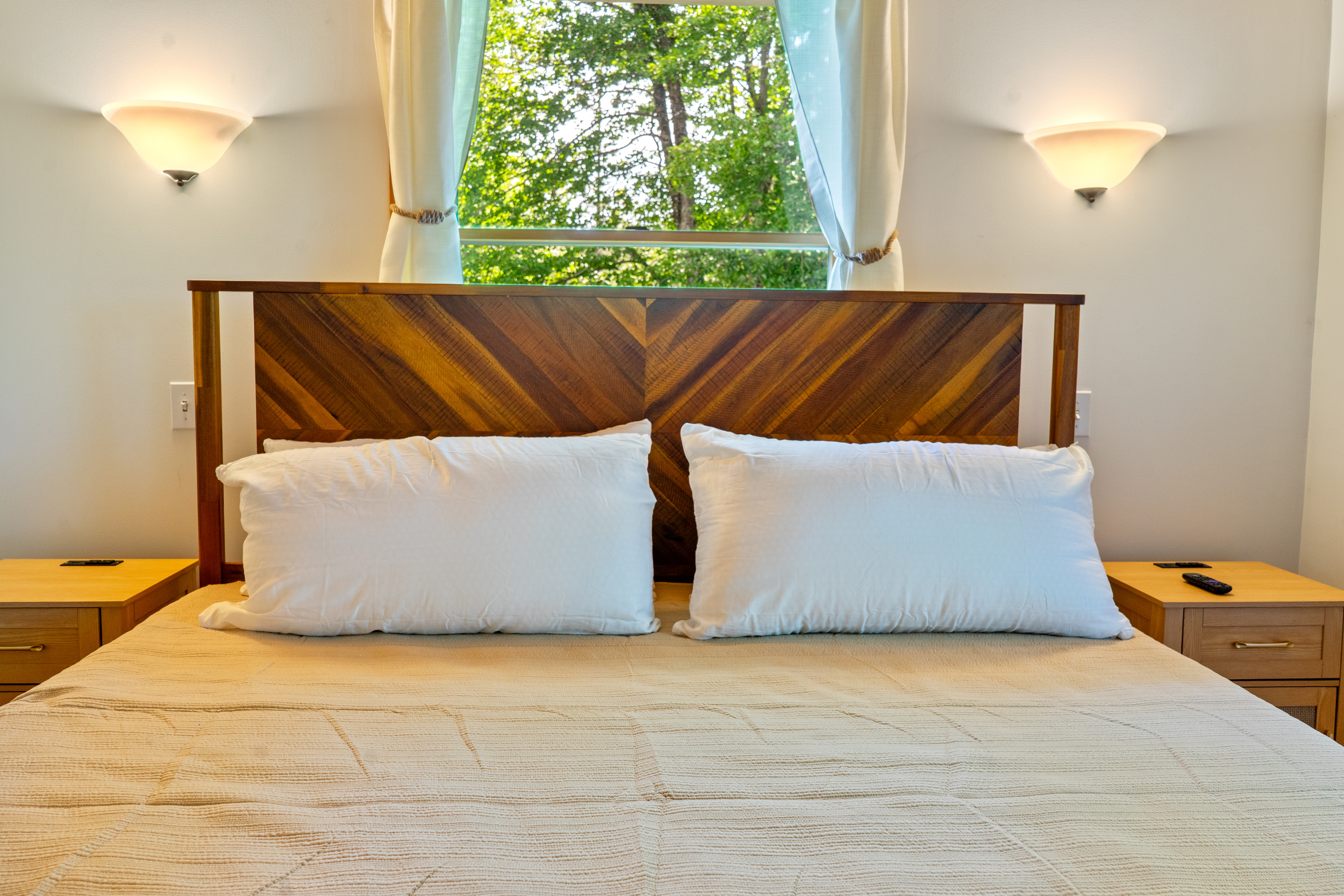 Unwind in the bright primary bedroom, anchored by a king bed with a signature chevron wood headboard. Positioned beneath a window, this symmetrical layout includes twin wooden nightstands and soft wall-mounted accent sconces.