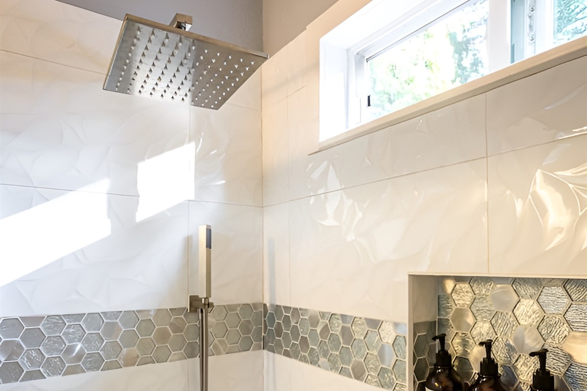- Sleek shower with rainfall head, handheld sprayer & contemporary hardware finishes
- White textured tiles accented with metallic hexagonal band for a designer look
- Built in recessed shelf keeps bath products organized & within easy reach for use
