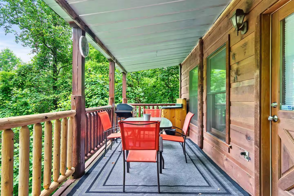 This inviting porch features a dining setup with bold red chairs, a glass table, and a grill, all under the shade of a covered roof, offering a tranquil and functional outdoor retreat.