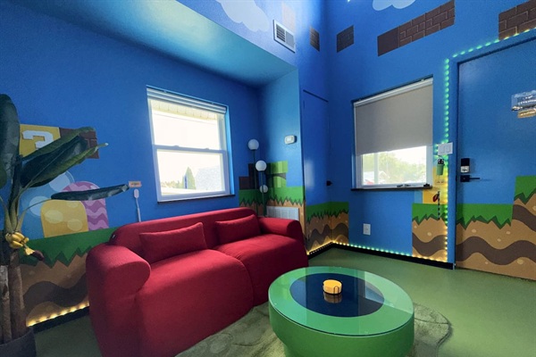 Relax on the bright red sofa in our Mario-themed living room at Mario's Mini Mansion, near Orlando, FL. Prop up your feet on the green pipe table amid hand-painted clouds, hills, and classic Super Mario decor. Ideal for cozy evenings!
