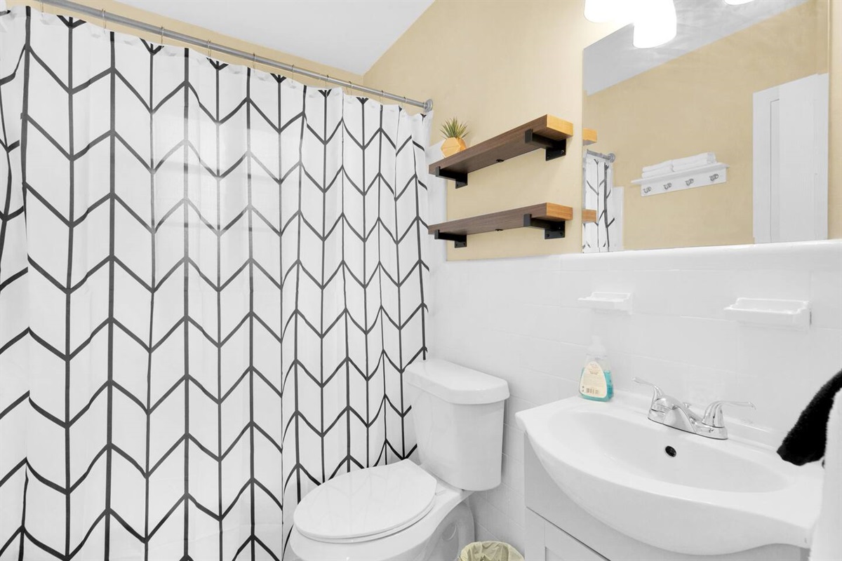 The bathroom has a full tub with a new versatile shower head. Large towels are stored under the sink while hand towels and wash cloths are stored above the towel hooks.