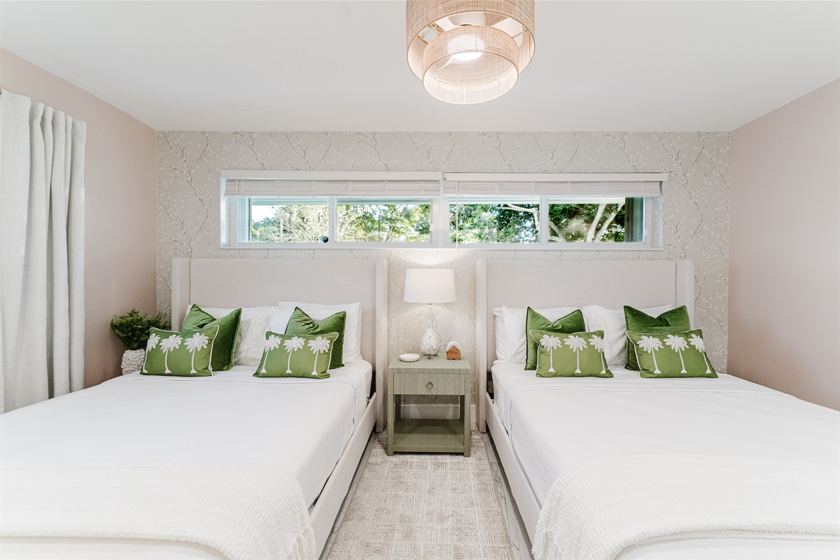 Bright, serene guest suite with two queen beds, hotel-quality linens, a wall-mounted smart TV, and ample storage. Features blackout curtains and blinds, a rattan ceiling fan, and a private en-suite bathroom—ideal for families or guests sharing space.