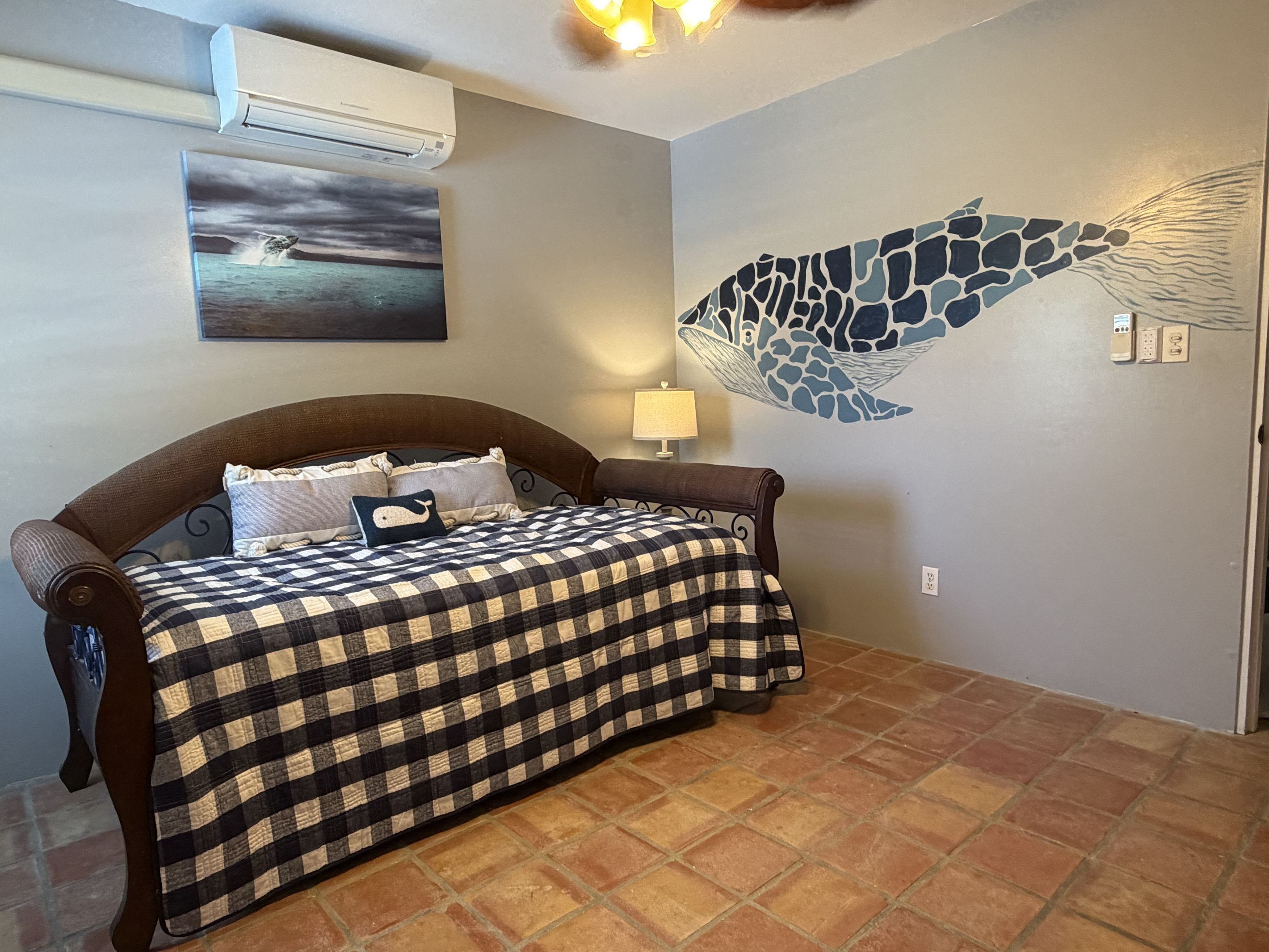 Features a versatile daybed with a pull-out trundle. Highlighted by a hand-painted mosaic whale mural and marine art, this room offers split-system AC, classic Saltillo tile floors, a tall distressed dresser, and a cozy bedside lamp with USB ports