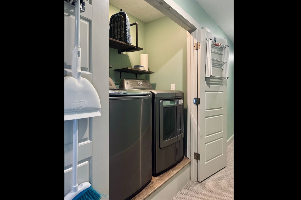 Convenient laundry closet featuring washer and dryer, ironing board, hanging rack, paper towels, and dryer sheets for everyday ease.