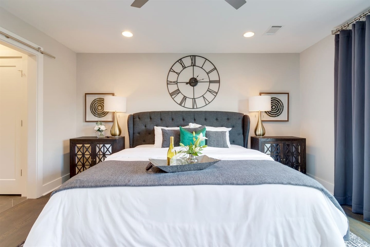 - Oversized tufted headboard anchors the space, paired with crisp white and charcoal bedding - Giant wall clock and matching lamps bring symmetry and bold visual appeal - Welcoming tray with wine and flowers adds an elegant, hotel style arrival