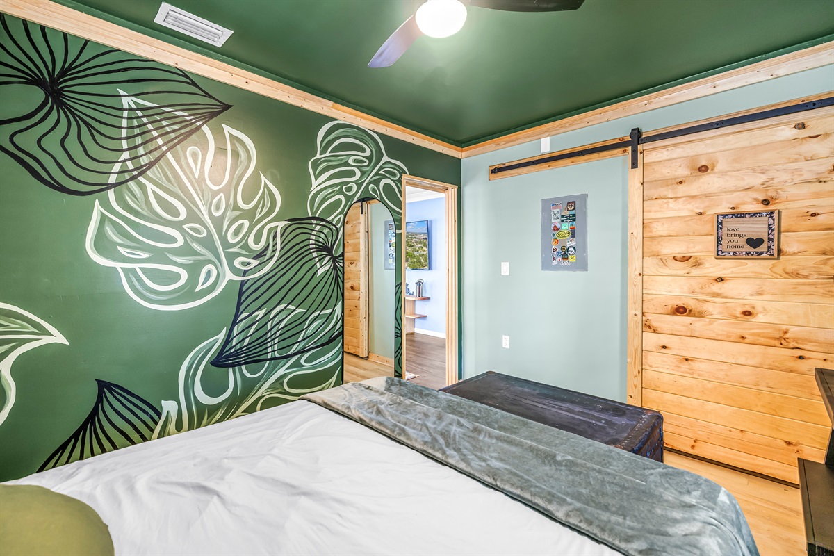 Cozy bedroom with a bold tropical accent wall, warm wood details, ceiling fan, and comfortable bedding for a relaxing stay.