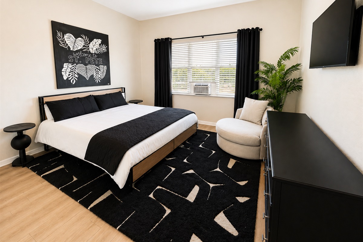 Stylish and cozy bedroom featuring a comfortable queen bed, soft linens, and modern décor. Enjoy natural light, a wall-mounted TV, and a relaxing reading chair—perfect for unwinding after a long day.