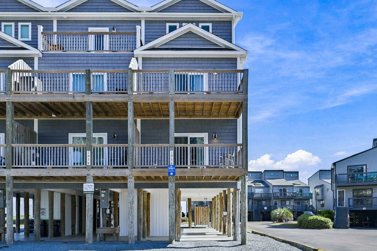 Exterior view showcasing the multi-level coastal townhouse with blue shaker siding, spacious private wooden balconies on each floor, and covered ground-level parking beneath the elevated structure.