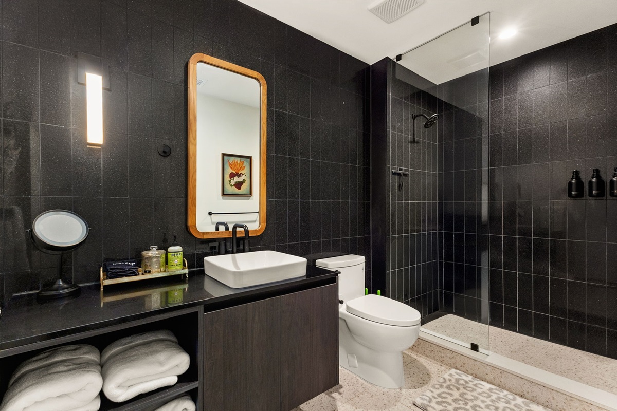 A luxurious bathroom featuring sleek black tile walls, a white vessel sink on a dark countertop, and a spacious walk-in shower with a glass partition, all complemented by a light-colored speckled floor and a modern, backlit mirror with a wooden frame.