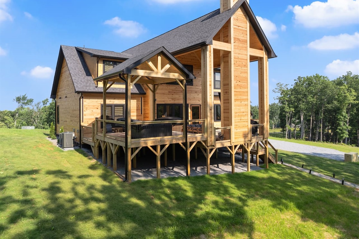 Admire the stunning craftsmanship of the cabin from this side angle, with its exposed beams, high peaks, and inviting hot tub area nestled under a charming pavilion.
