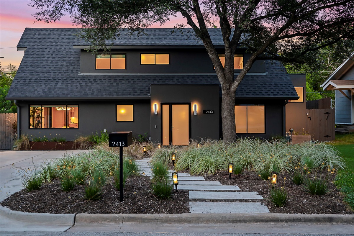A contemporary two-story home with a dark gray facade, a minimalist front yard with a stepping-stone path leading to the front door, and subtle landscape lighting, all captured during a vibrant sunset.