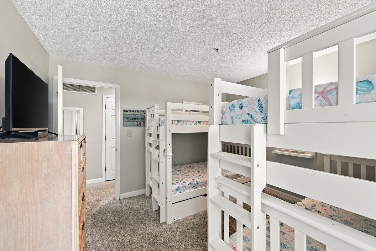 Spacious bunk room configured with dual white wooden bunk bed sets, 1 trundle, pastel sea life linens, flat-screen TV mounted above a weathered wood dresser, and a seamless hallway entry.   