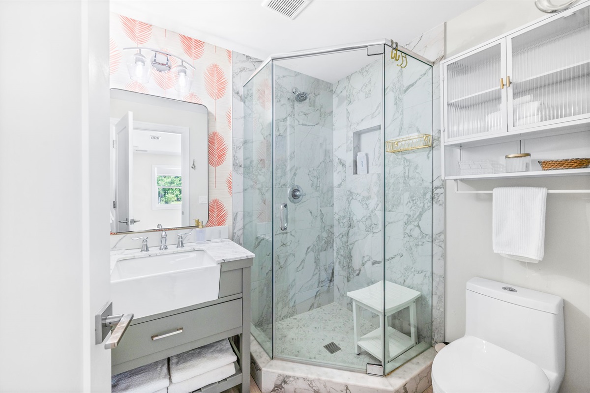 Here's the full bathroom in the downstairs master—compact but beautifully designed. A glass shower, coral palm leaf wallpaper, and a white sink with a glass storage cabinet above the toilet make this space both functional and stylish.