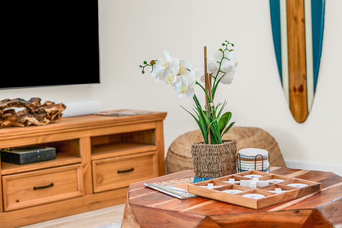 The bright and airy living room features a modern leather sectional, warm wood accents, and lush greenery views through large picture windows. Stylish touches like the surfboard décor and geometric coffee table create a relaxed coastal vibe.