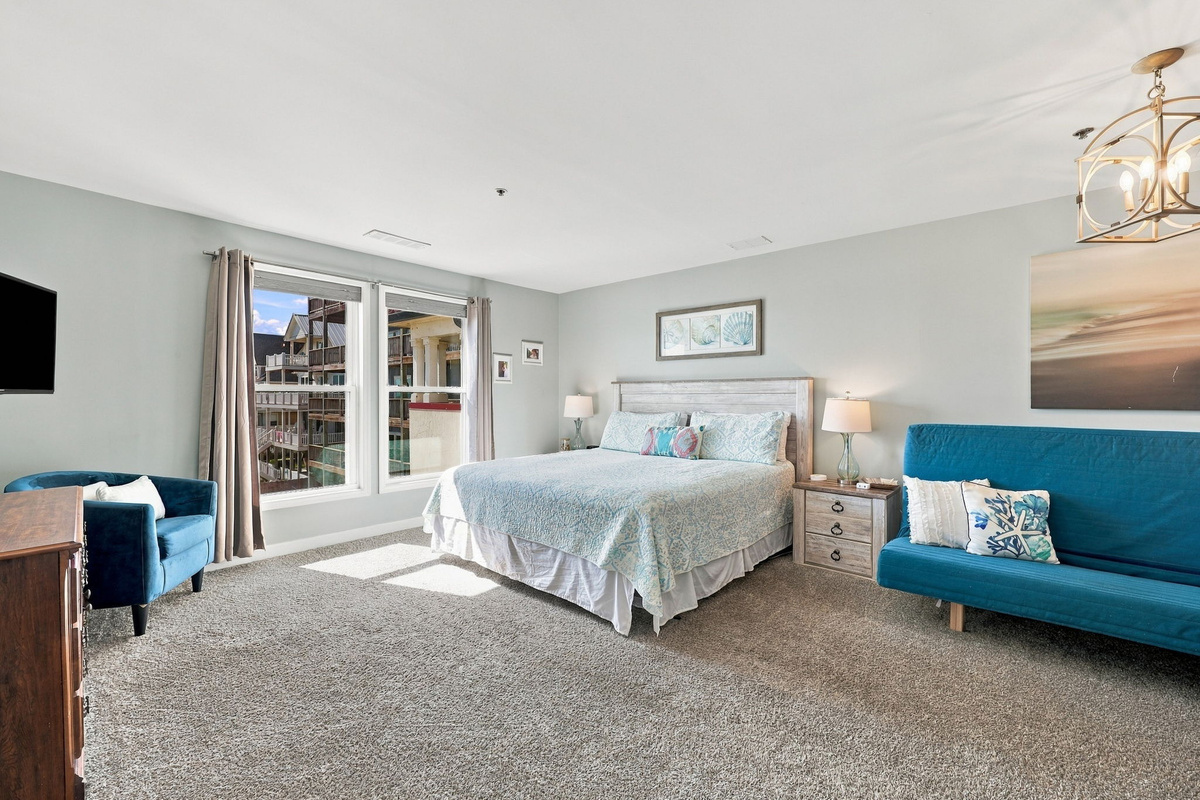 Expansive primary suite featuring a king bed with weathered wood headboard, vibrant blue futon, cozy armchair nook, lantern chandelier, and large windows with coastal community vistas.   