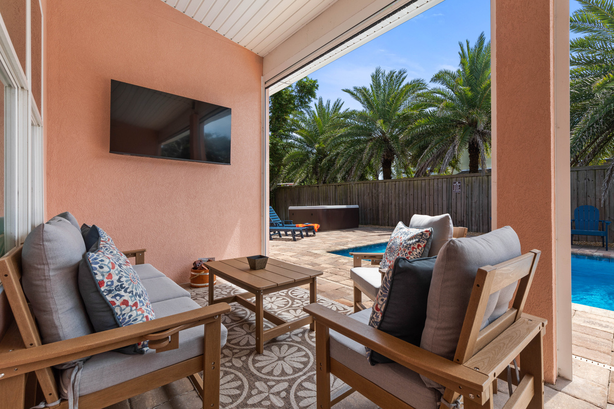 Covered lanai seating faces a wall-mounted TV with the pool just beyond, so you can watch the game while kids swim.