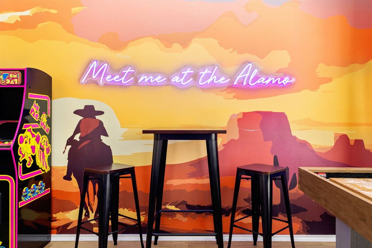 Fun and vibrant game room corner featuring a custom Texas-themed mural with neon “Meet me at the Alamo” sign, arcade game, and pub-style seating—perfect for friendly competitions, cocktails, and unforgettable San Antonio nights.
