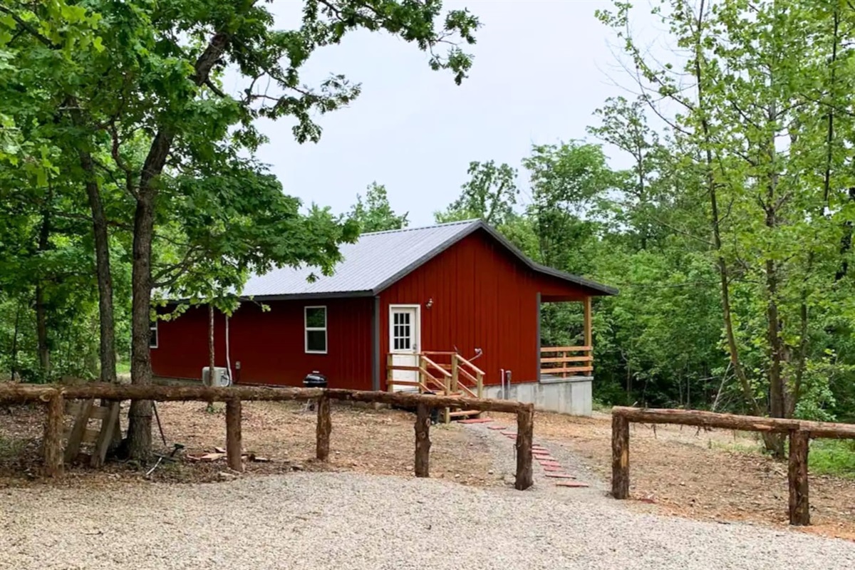 Misty Oak Lodge sits quietly among the trees with rustic fencing, a gravel drive, and a warm red exterior. The covered porch and wooded surroundings make it the perfect private escape for peaceful mornings, evening grilling.