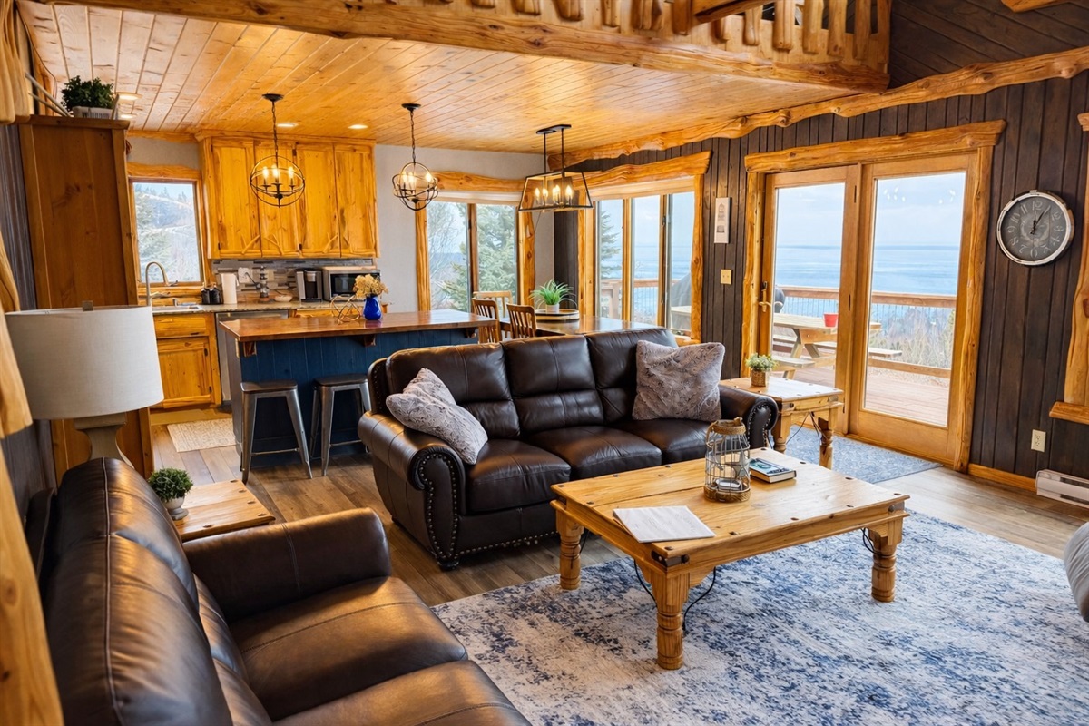 Cozy seating area with a comfy couch and loveseat around the coffee table, with Lake Superior peeking through the windows. Everything flows together, with the kitchen and dining space in the background.
