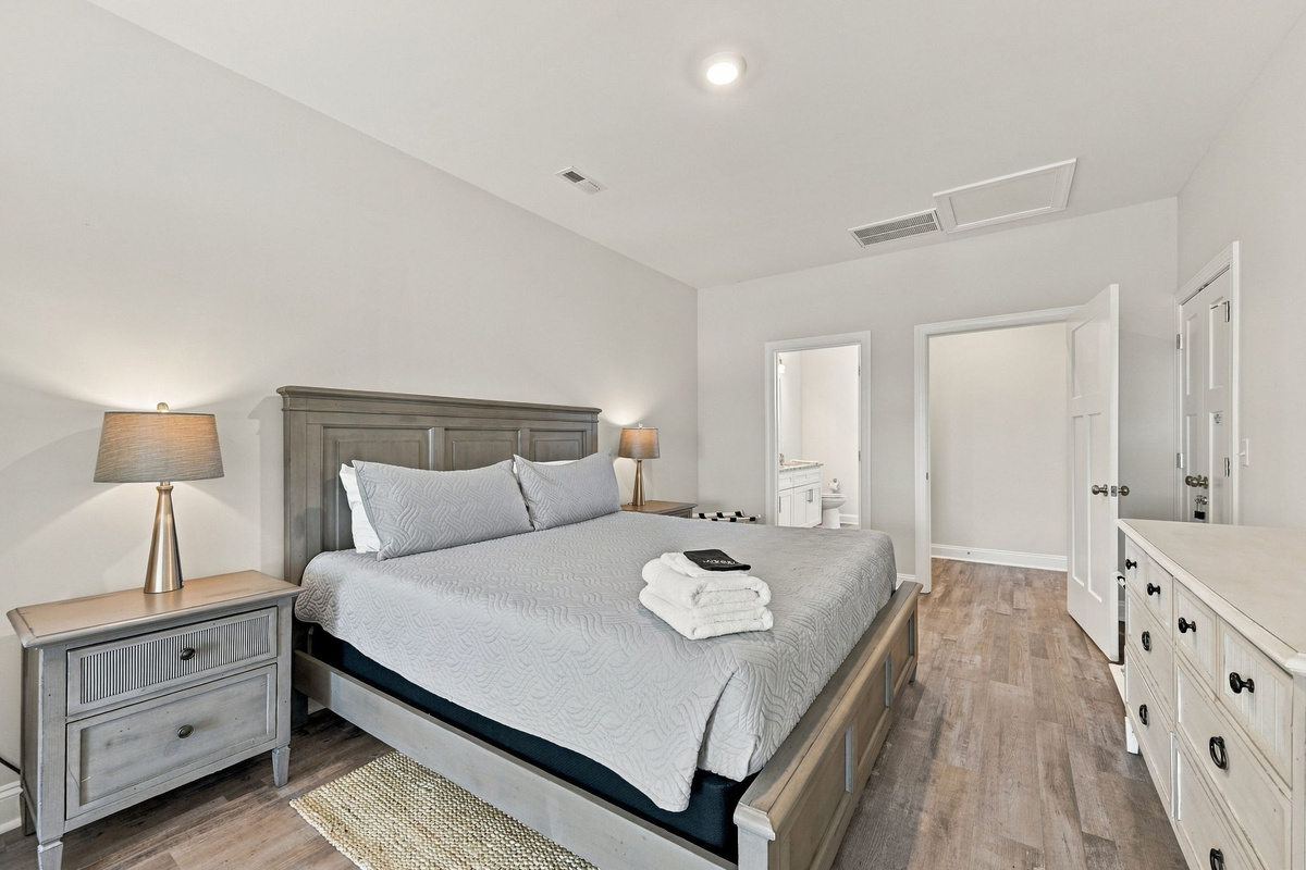 Spacious primary suite featuring a king-sized bed with a weathered wood panel headboard, matching nightstands with elegant lamps, a white multi-drawer dresser, and direct access to an en-suite bathroom.   
