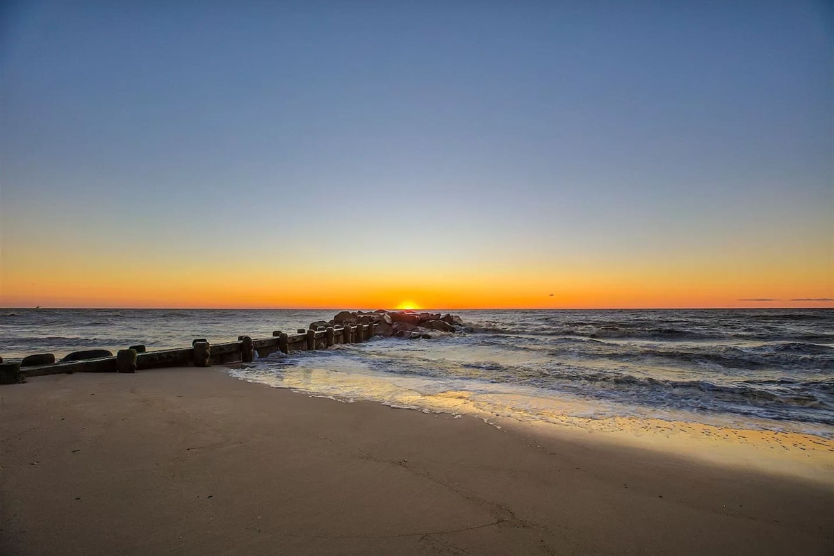 Capture a serene moment on the beach as the sun sets, painting the sky in warm shades of orange and gold. The gentle waves wash over the sand near a weathered wooden breakwater, offering a peaceful backdrop for your coastal getaway.