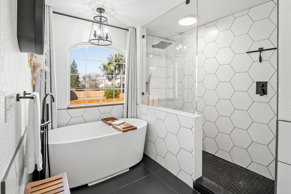 Spa-like primary bathroom featuring a freestanding soaking tub beneath a picture window and a spacious walk-in shower with striking hex tile. Modern black fixtures and elegant lighting create a bright.
