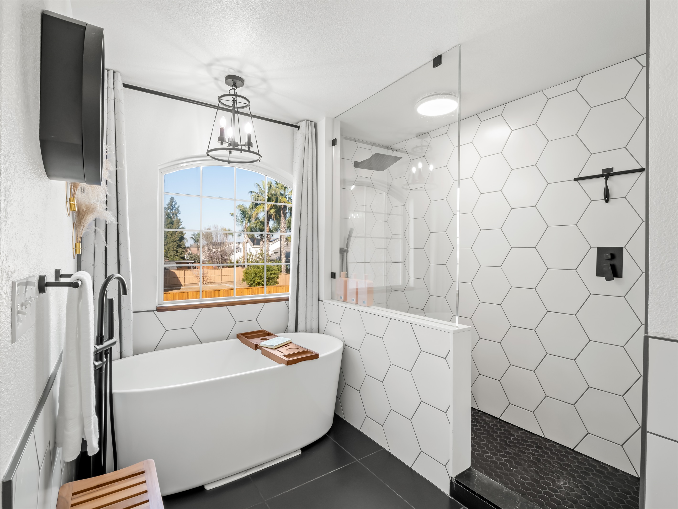 Spa-like primary bathroom featuring a freestanding soaking tub beneath a picture window and a spacious walk-in shower with striking hex tile. Modern black fixtures and elegant lighting create a bright.
