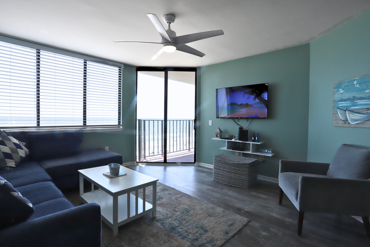 A chic TV lounge with flat tv screen , adaptable white and blue furnishings, large windows that invite abundant natural light, and a balcony showcasing stunning coastal views—ideal for unwinding and entertaining.