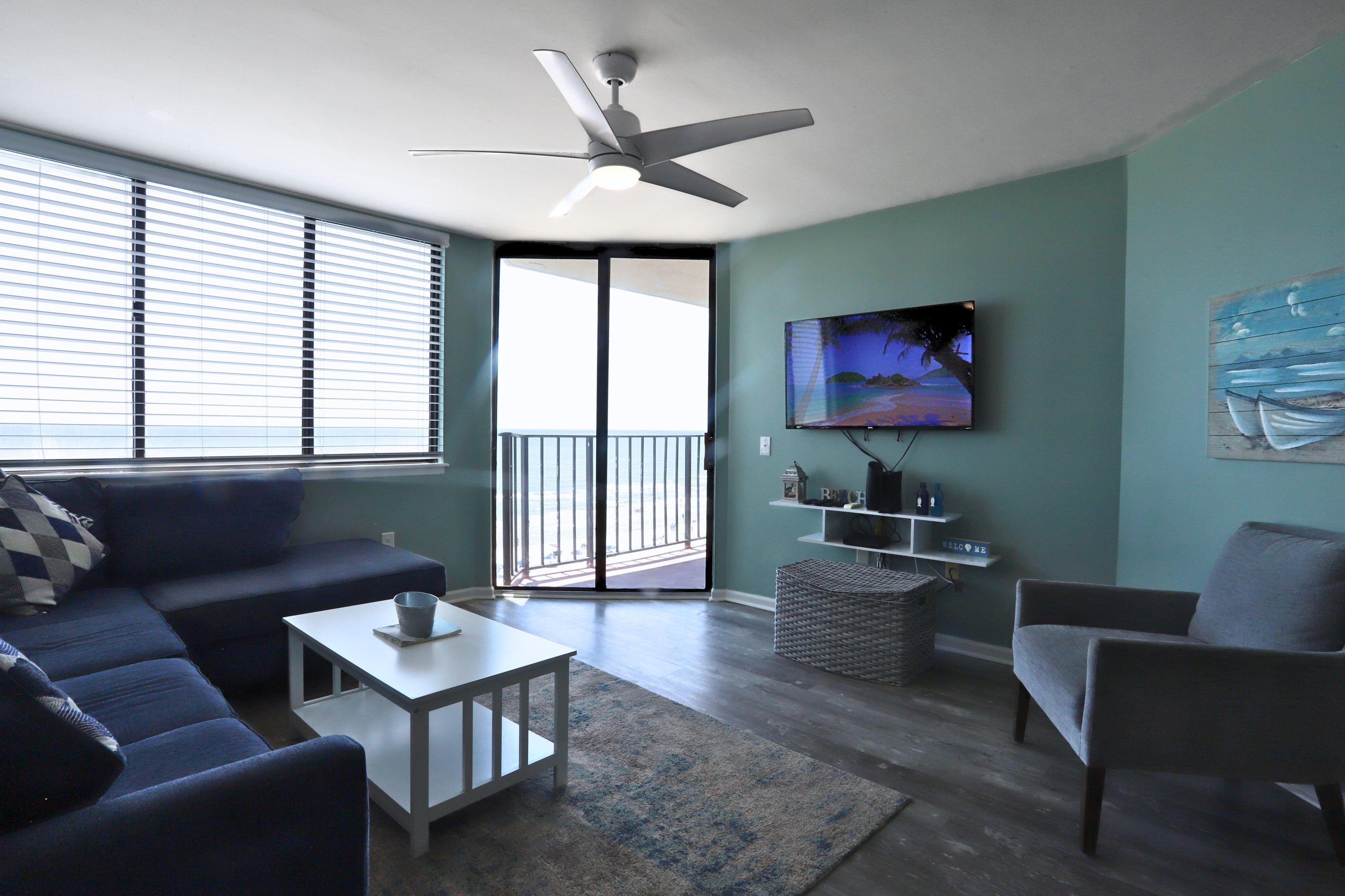 A chic TV lounge with flat tv screen , adaptable white and blue furnishings, large windows that invite abundant natural light, and a balcony showcasing stunning coastal views—ideal for unwinding and entertaining.