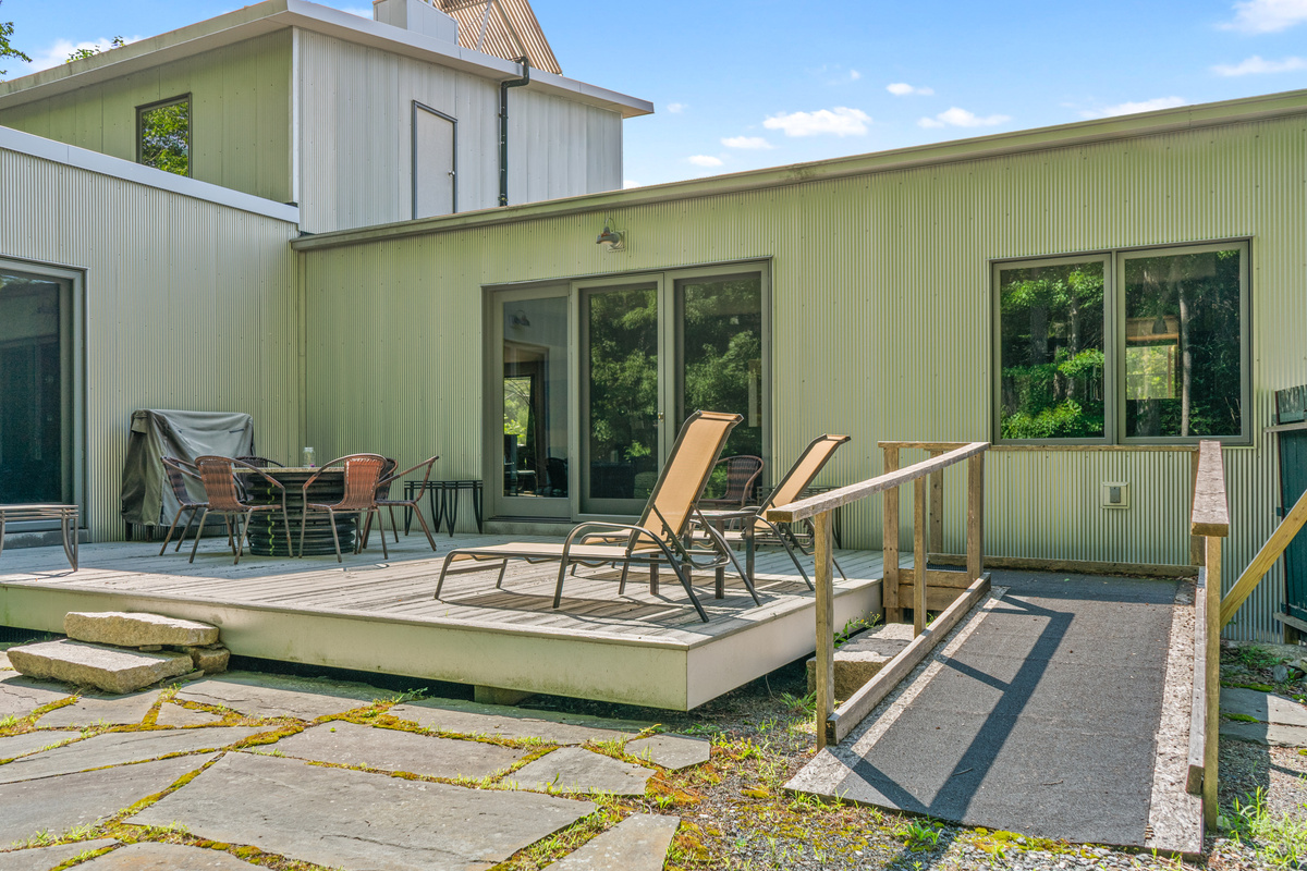 The expansive 33-foot outdoor deck at "The Crag." Features stone hardscaping, a step-free accessibility ramp, sun loungers, dining space, and seamless glass door entries—all set right along the boundary of Acadia National Park.