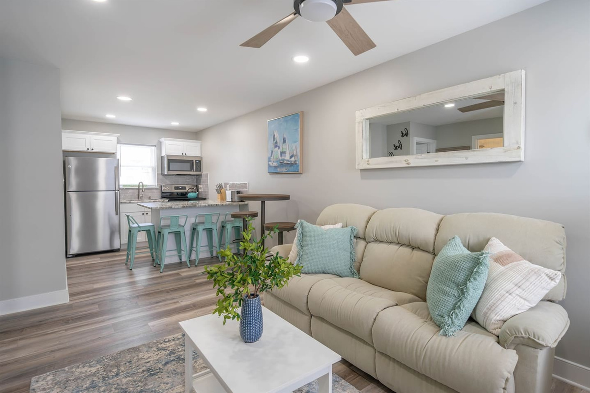 Open, airy living area flowing into the kitchen, featuring plush seating, ceiling fan, and soft coastal styling. The layout encourages relaxed lounging, easy conversation, and casual meals, making it a comfortable hub for unwinding throughout the sta
