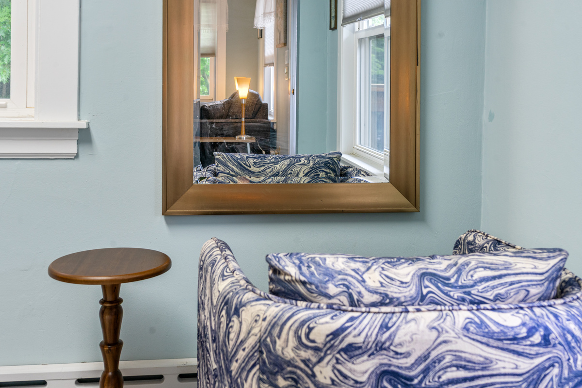 A close-up view highlights the compass den vanity layout. Complete with a blue-and-white patterned accent chair, a round wooden pedestal table, and a large gold-framed wall mirror, this arrangement offers an additional area to get ready.