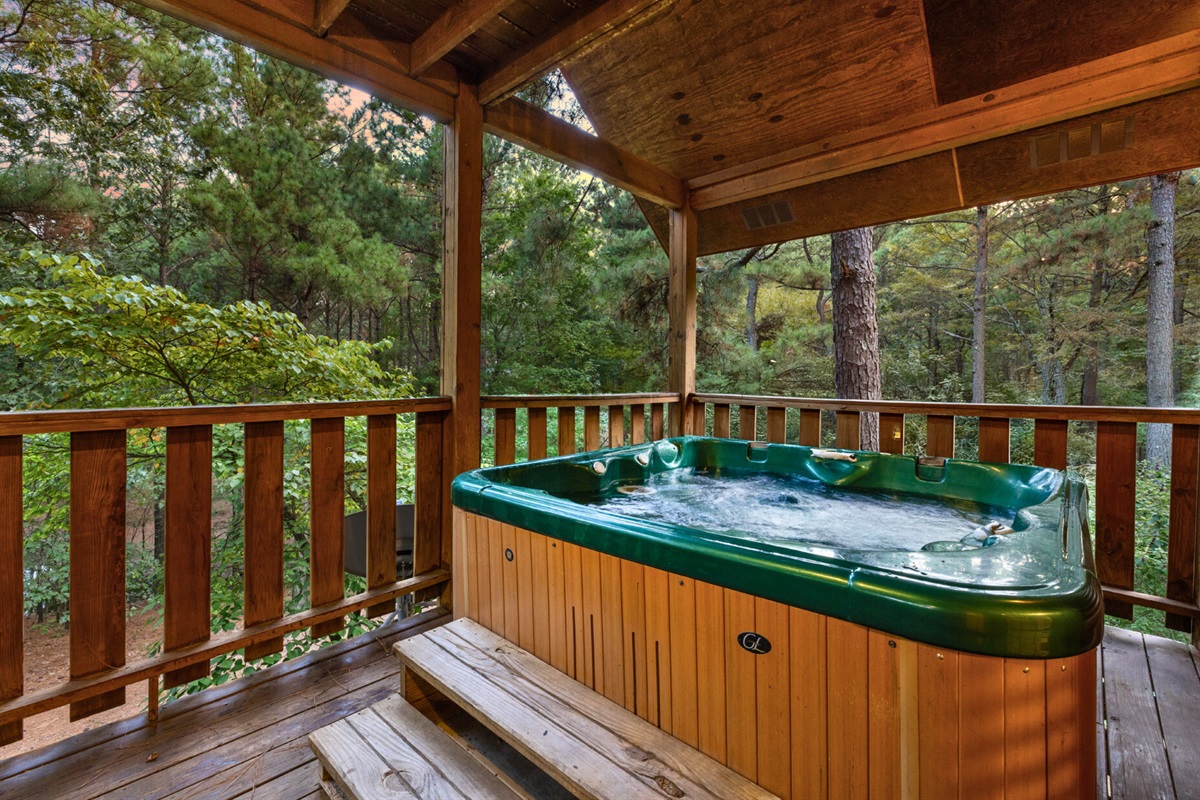 Relax in the hot tub on a covered deck surrounded by tall pines.