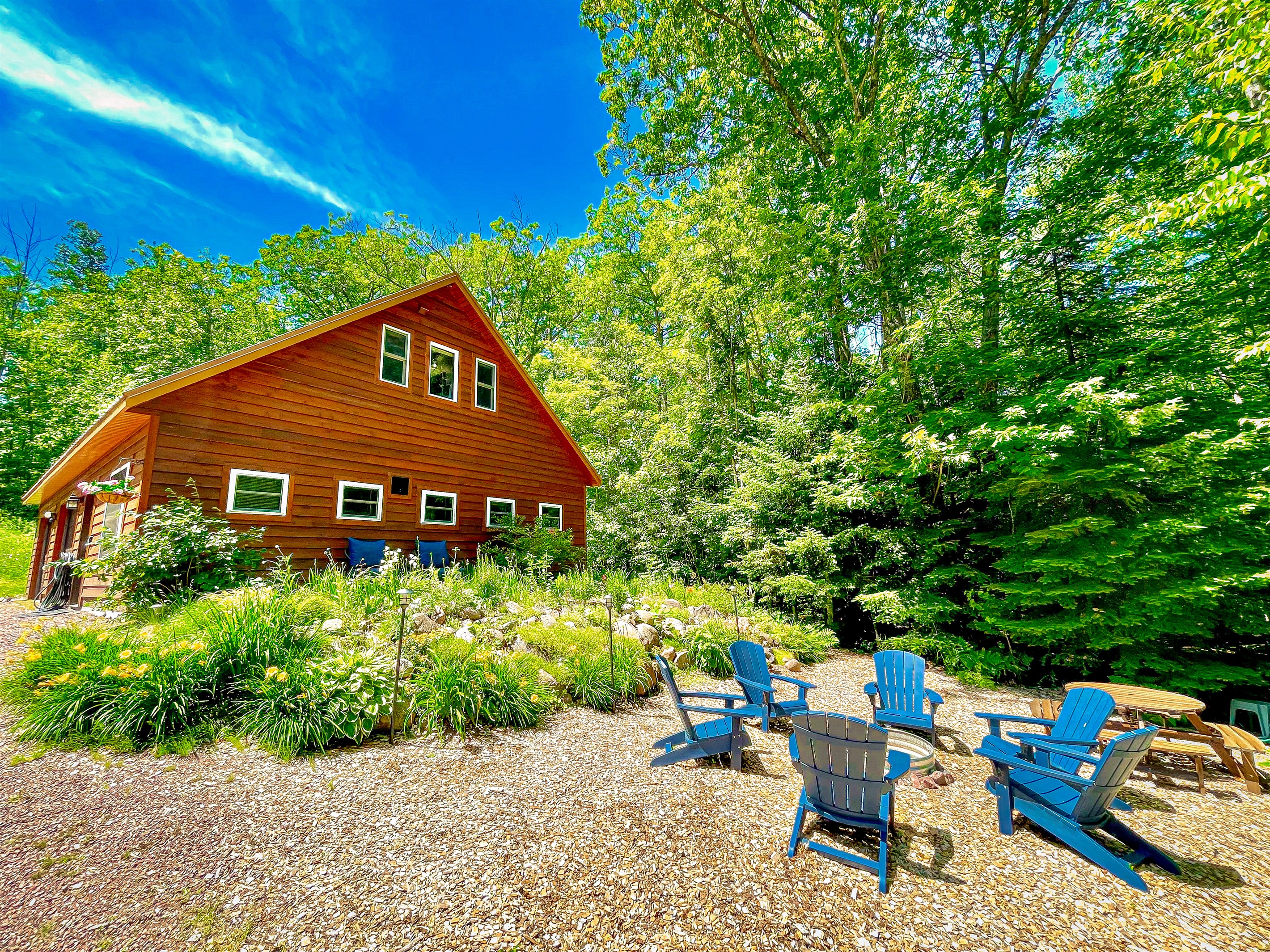 VIew of Rock Garden driving up to property from the road- in the summer  Showing perennial garden fire pit with provided wood and picnic table 