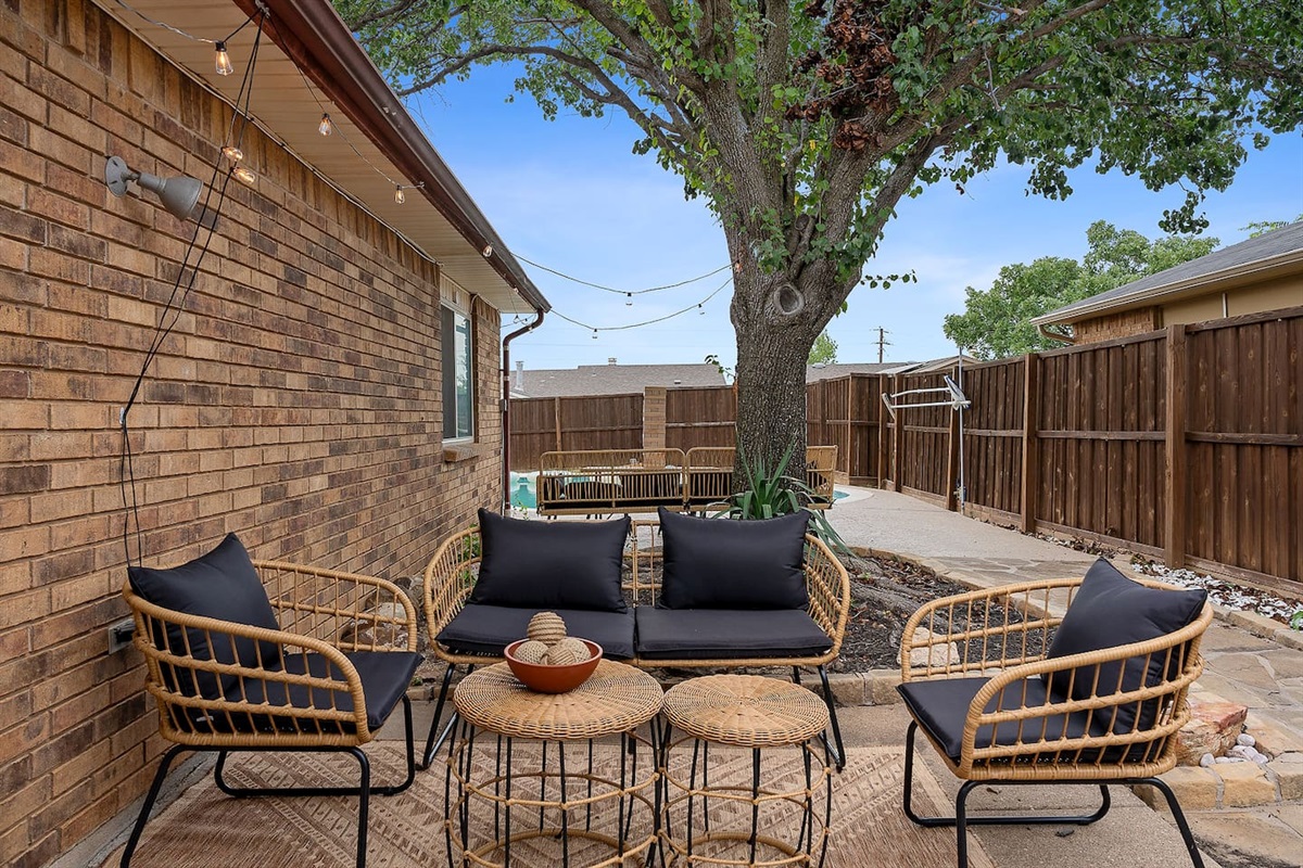 Discover this charming outdoor patio space with chic wicker chairs beneath a canopy of lights and a shady tree.