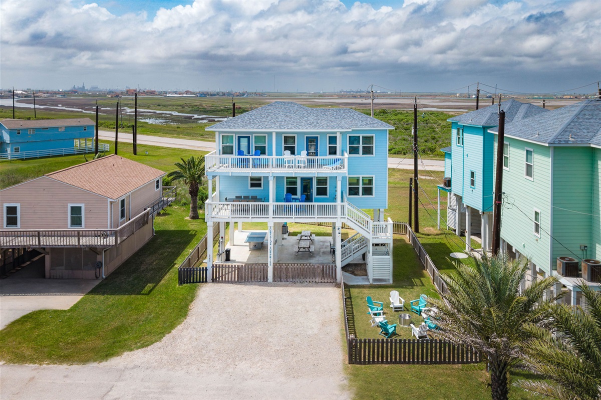 Daytime exterior — classic stilted Surfside beach house. Big blue sky, big blue house, your week's home.