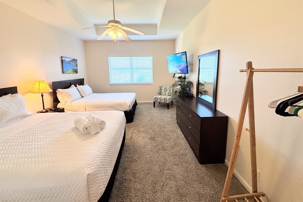 Spacious bedroom featuring two comfortable beds, TV, ceiling fan, dresser storage, and a convenient luggage rack.