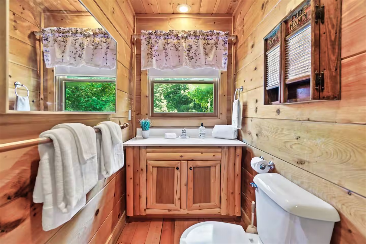 This charming bathroom features a wooden vanity with modern fixtures, fluffy white towels, and a sunlit window with floral curtains that offer a serene view of the outdoors.